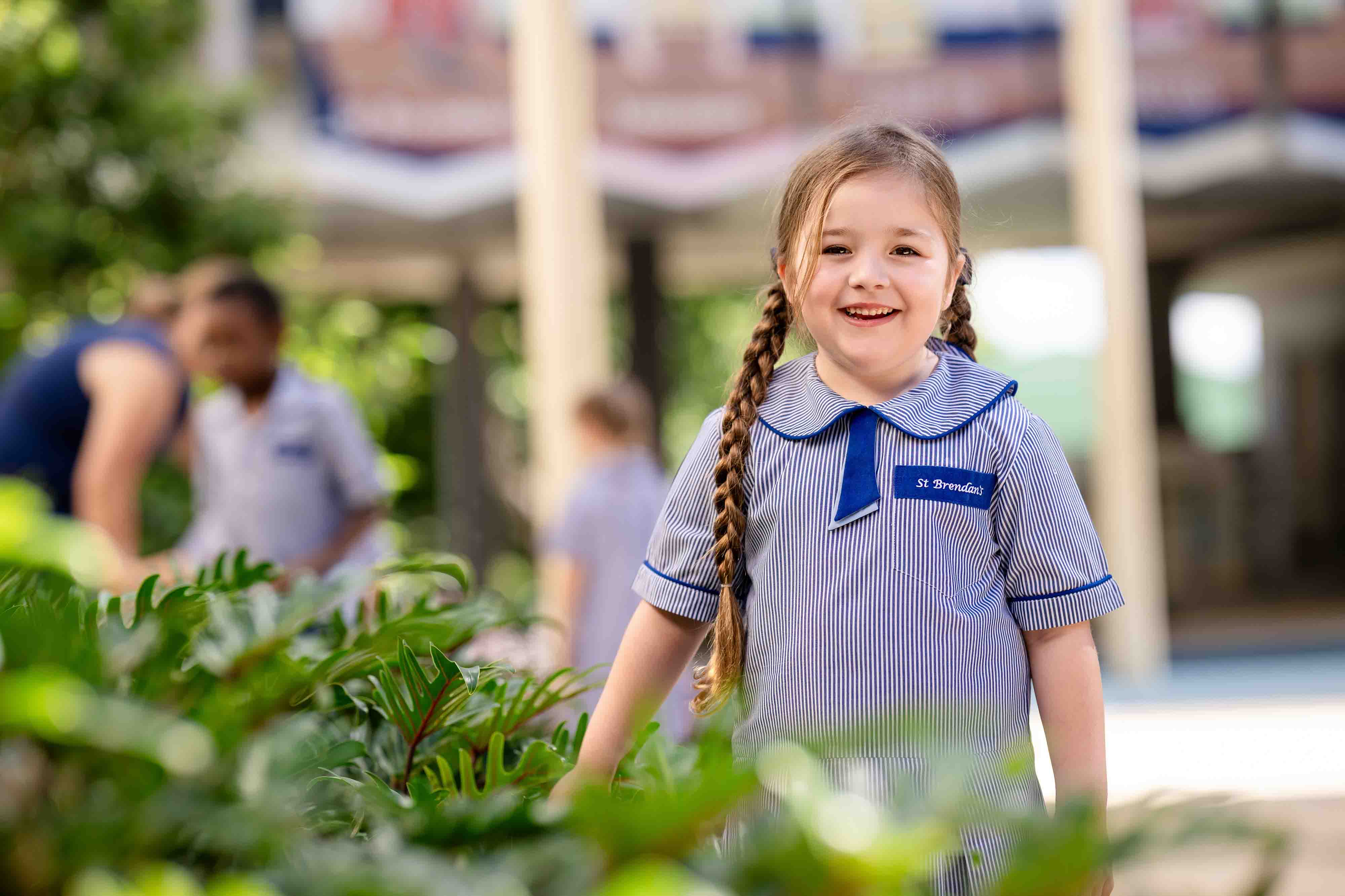 Student smiling in school uniform standing among greenery with classmates in background during outdoor learning activity