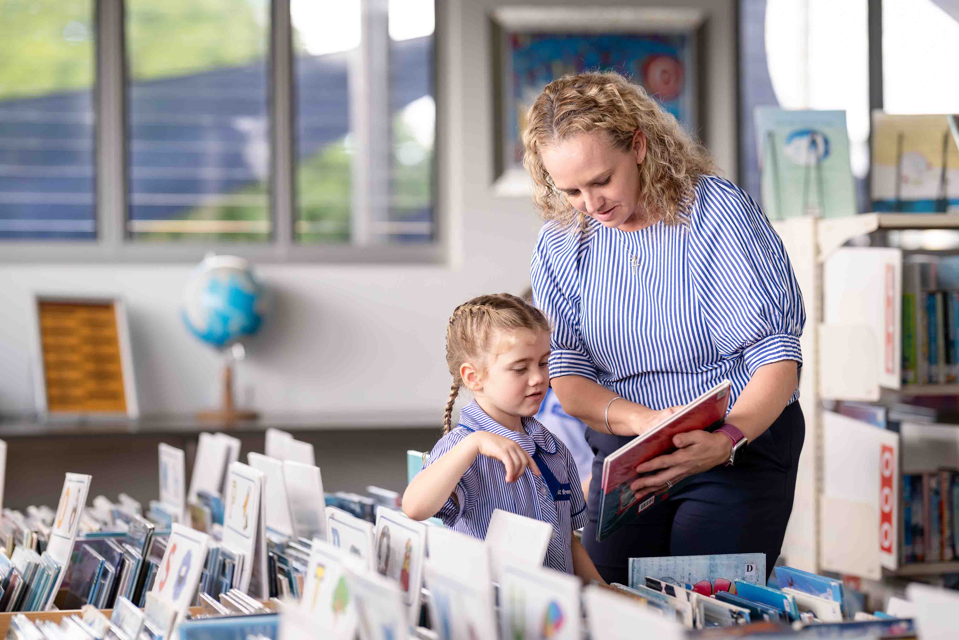 Teacher supporting young student while selecting and reading book together in school library