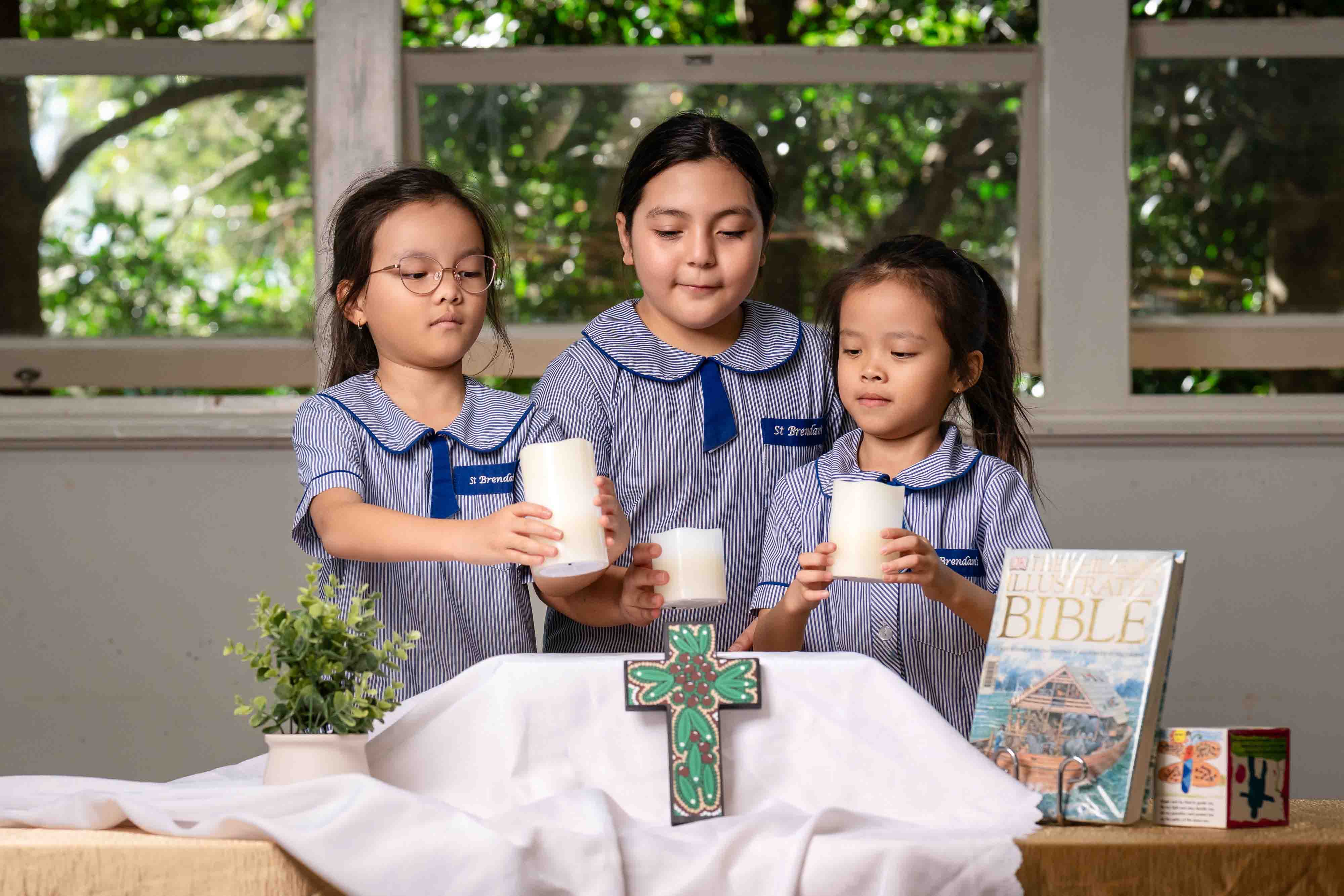 Students participating in prayer activity holding candles around table with cross and Bible in classroom setting