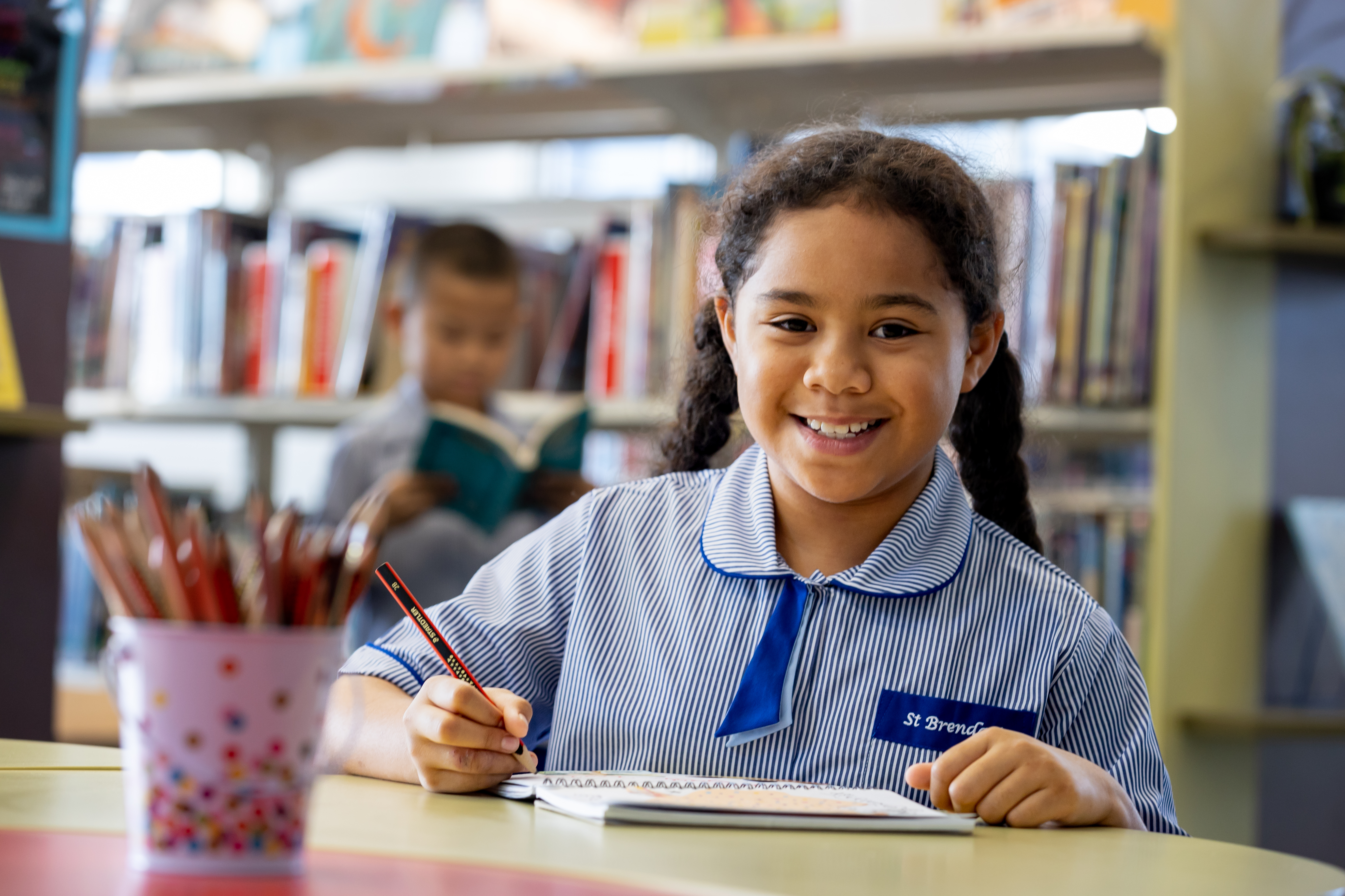 Student smiling while writing in notebook in classroom with bookshelves and classmate reading in background